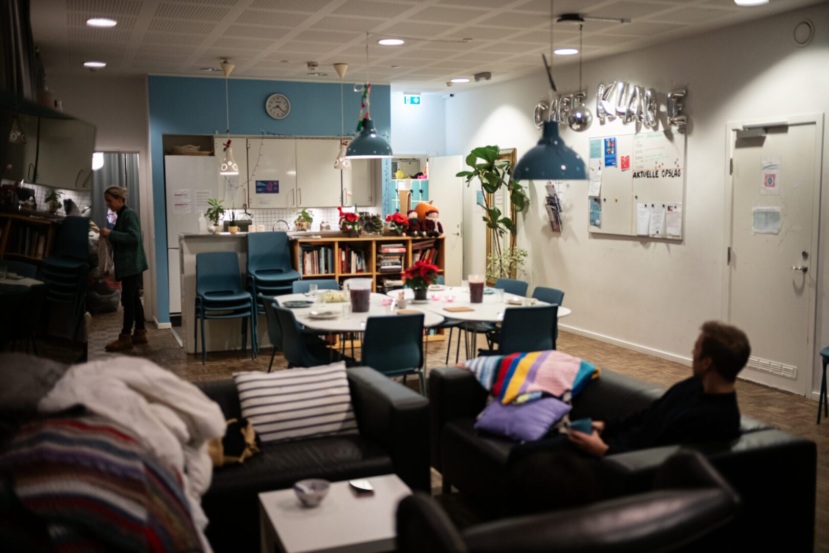 Hyggeligt fællesrum med borde, stole og sofaer. En person ser TV, mens en anden står ved køkkenet.